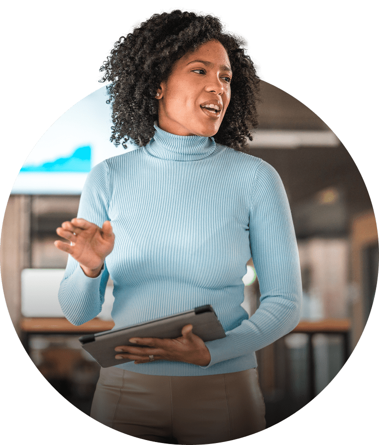 A woman in a blue polo necked jumper holds a tablet computer whilst she presents in front of a room.